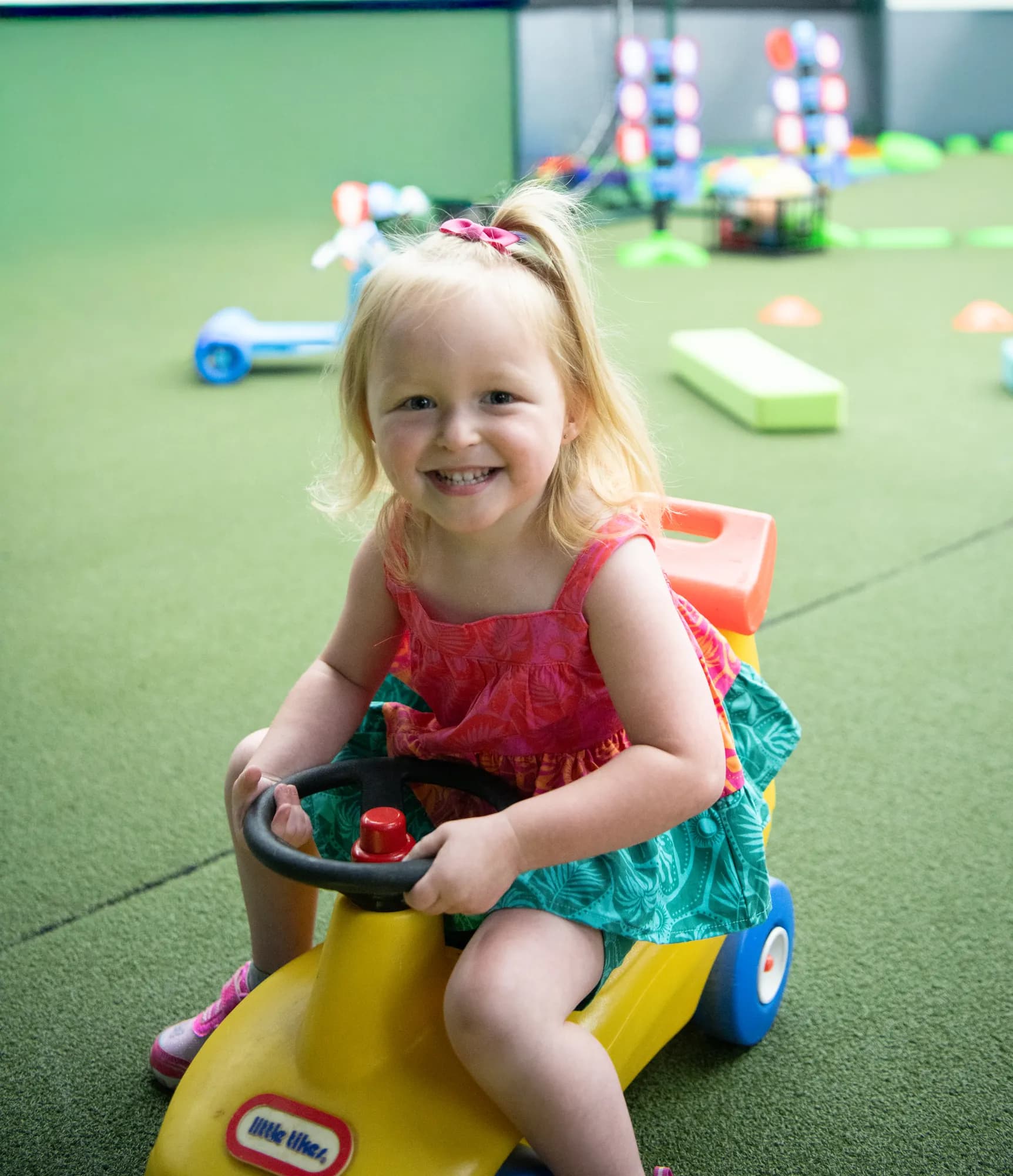 A smiling toddler riding a yellow ride-on car inside the CHAMP Academy space at The Hitter's Edge